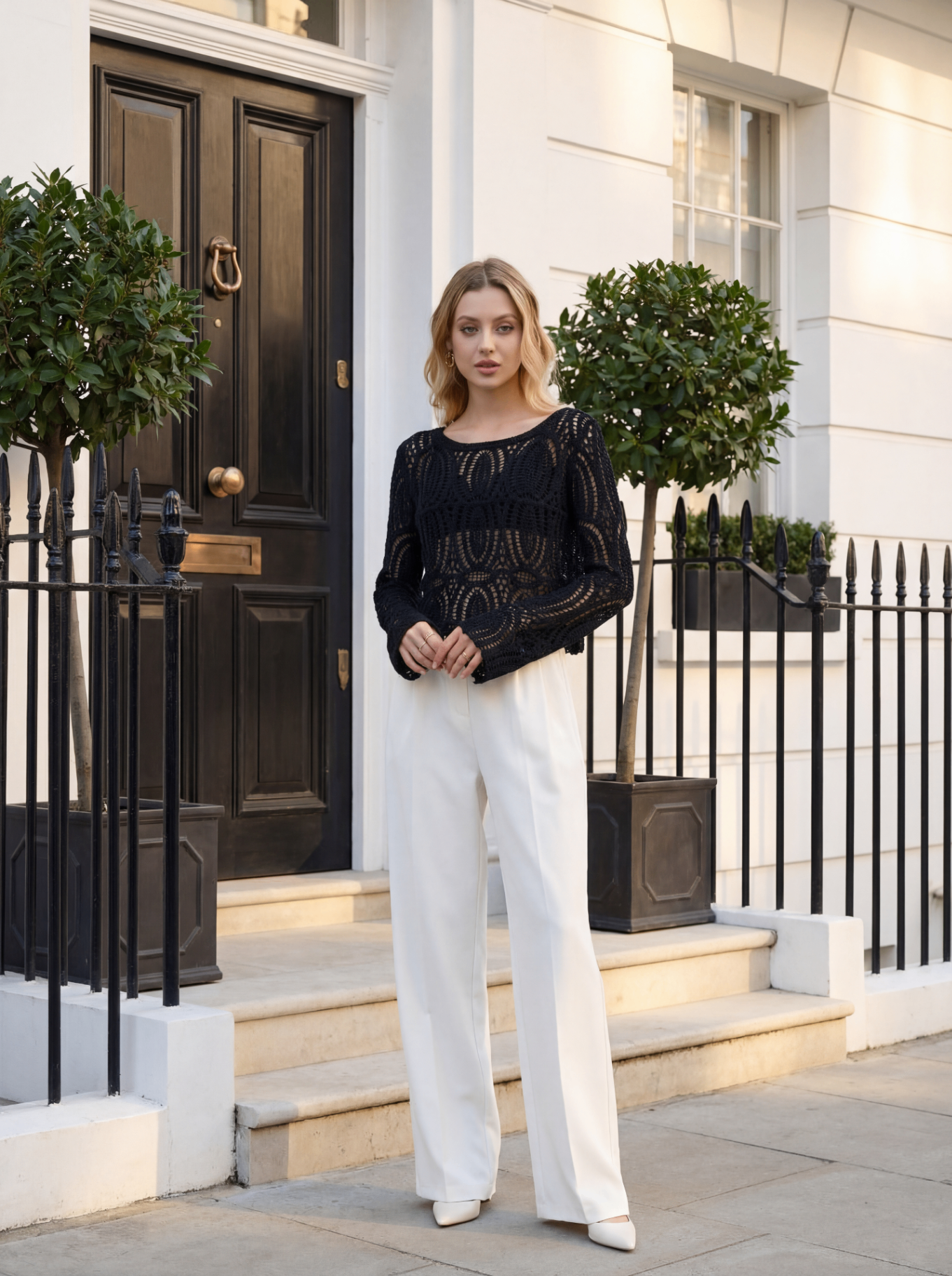 Девушка в чёрном ажурном свитере и белых брюках у входа в дорогой дом с чёрной дверью